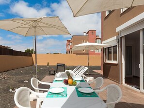 Uma área de refeições ao ar livre com uma mesa posta para quatro pessoas, dois guarda-sóis e vista para edifícios ao fundo.