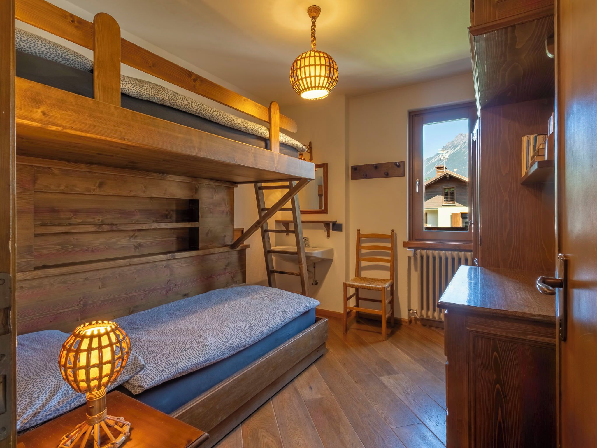 A bunk bed room with a ladder, a chair, a wooden cabinet, and a window with a view.