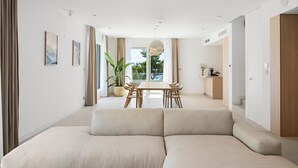 Un salon moderne avec un canapé d’angle, une table à manger avec des chaises, une grande plante en pot et une vue sur l’extérieur.