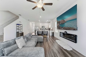 A modern living room with a gray sectional sofa, a ceiling fan, a kitchen area with a bar, and a large ocean view painting. - Covey Luxe Modern Getaway (Davenport)