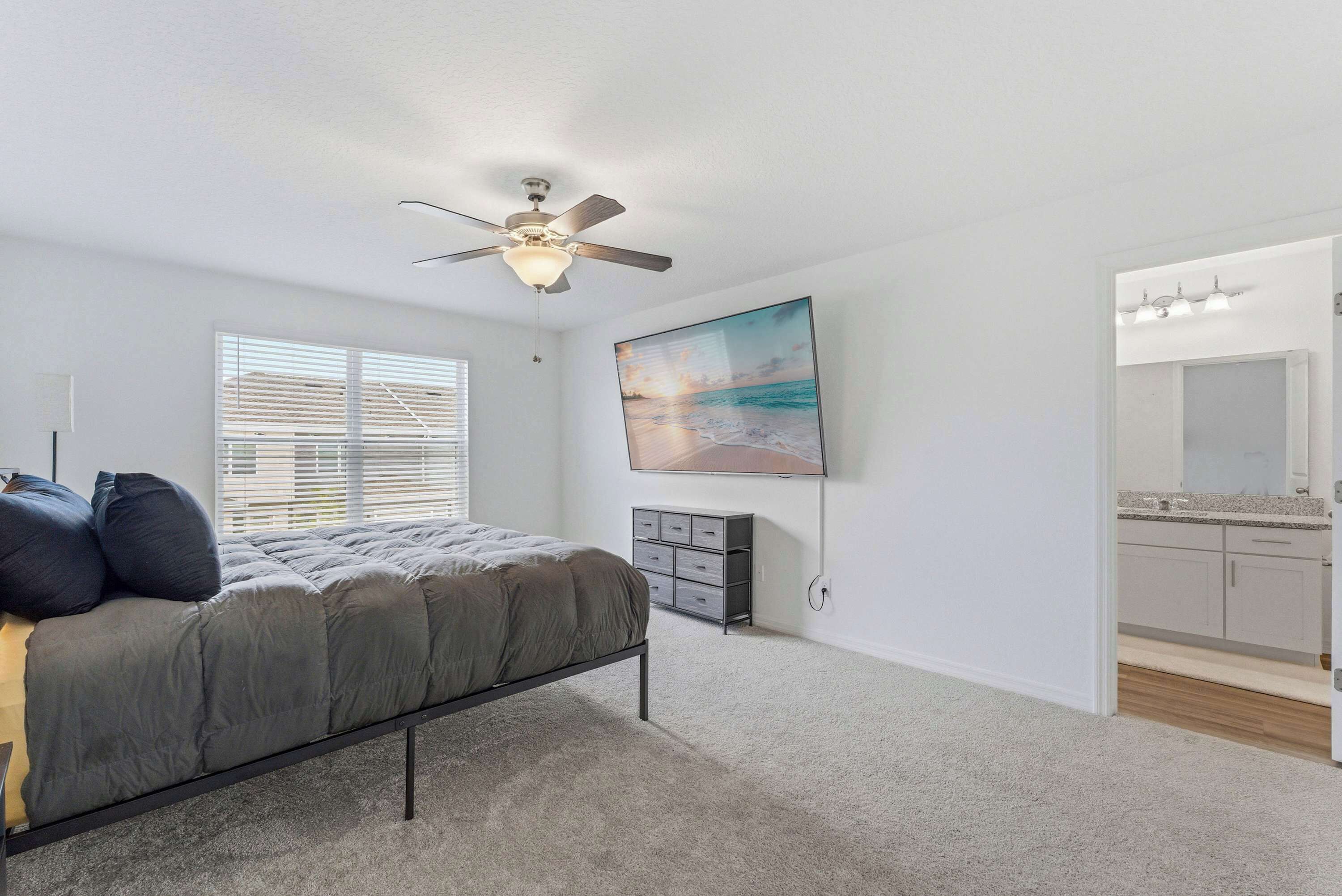 A bedroom with a bed, ceiling fan, artwork, dresser, and a bathroom with a mirror and countertop.