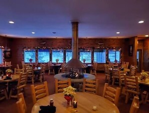 A restaurant with round tables, wooden chairs, a central chimney, and large windows.