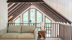 Dormitorio tipo loft con techo inclinado, un sofá, una lámpara y vistas a árboles a través de amplios ventanales.