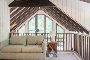 Ein Schlafzimmer im Loft-Stil mit schrägem Dach, einer Couch, einer Lampe und Blick auf Bäume durch große Fenster.