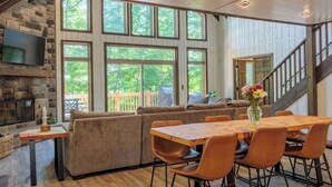 Un amplio salón con una mesa de comedor de madera, asientos acolchados, una chimenea de piedra y grandes ventanas que ofrecen vistas al exterior.