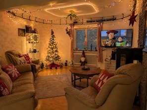 A cozy living room with a Christmas tree, comfortable seating, a fireplace, and a TV displaying a cartoon.