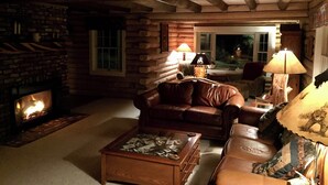 Interior acolhedor de uma cabana de madeira, com lareira, móveis de couro e vista para o exterior.