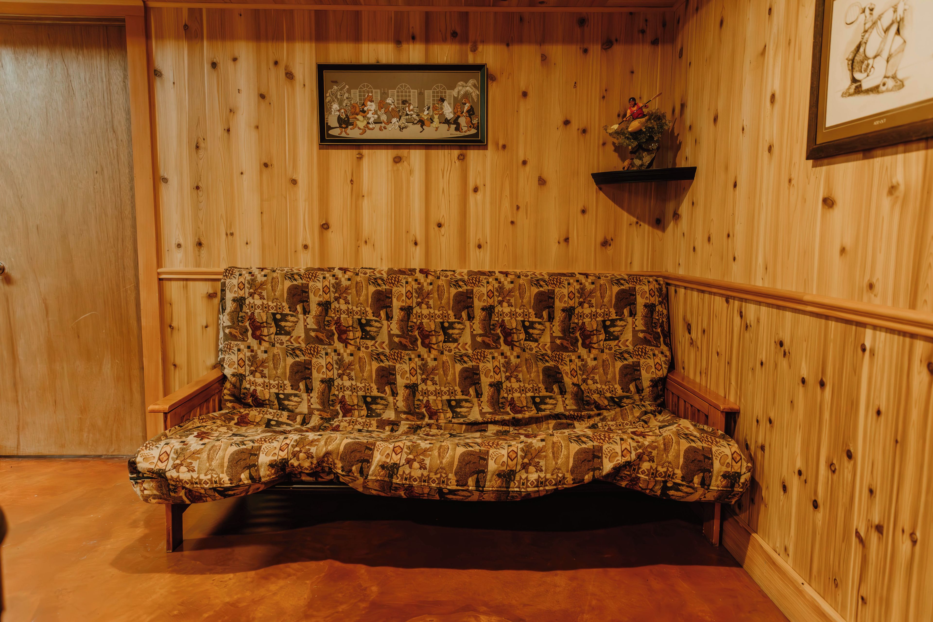 A wooden-paneled room with a patterned sofa, a framed picture on the wall, and a small shelf with a plant.