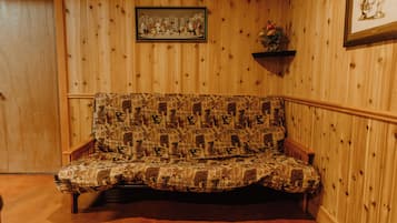 A wooden-paneled room with a patterned sofa, a framed picture on the wall, and a small shelf with a plant.