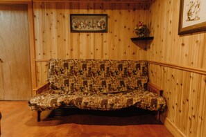 A wooden-paneled room with a patterned sofa, a framed picture on the wall, and a small shelf with a plant.