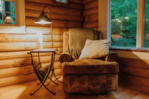 Acogedora habitación tipo cabaña de madera, con una silla, una mesa con lámpara y ventana con vista a árboles.