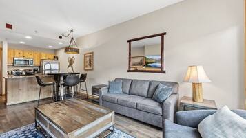 Un salon moderne avec un canapé gris, une table basse en bois et un coin repas comprenant des chaises, avec une cuisine en arrière-plan.