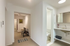 Corredor que dá para um banheiro com vaso sanitário, pia e espelho. Há um quadro na parede e uma pequena mesa com uma cadeira.