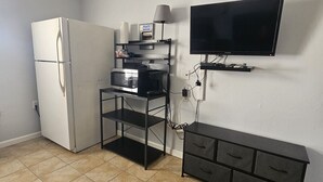 Una kitchenette equipada con refrigerador, microondas y estufa, un televisor montado en la pared, y un mueble pequeño con cajones.