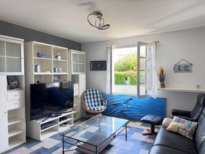A living room with a TV, glass coffee table, seating area, and a window with a view of greenery. - The Lighthouse Villa (Saint-Denis-d'Oléron)