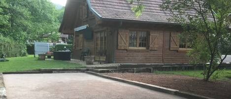 Uma cabana de madeira com telhado inclinado, um caminho de acesso pavimentado e uma área gramada com um banco.