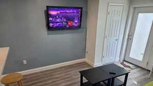 A living room with a flat-screen TV mounted on the wall, a black coffee table, a green sofa, and wooden stools. - 2 Bedroom Condo Less Than A Mile To Rehoboth & Dewey Beaches (Rehoboth Beach)