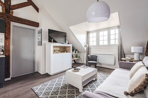 Un dormitorio estilo loft con techo inclinado, un ventanal grande, una cama blanca, una mesita de centro blanca y un televisor montado en la pared.