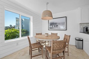 Un coin repas avec une table ronde, des chaises en osier, un luminaire suspendu, un tableau encadré au mur et une vue sur les bâtiments à travers de grandes fenêtres.