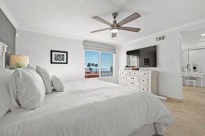 Ein Schlafzimmer mit Bett, Deckenventilator, Schminktisch, Fernseher und einem durchgehenden Türrahmen einsehbarer Badezimmer.