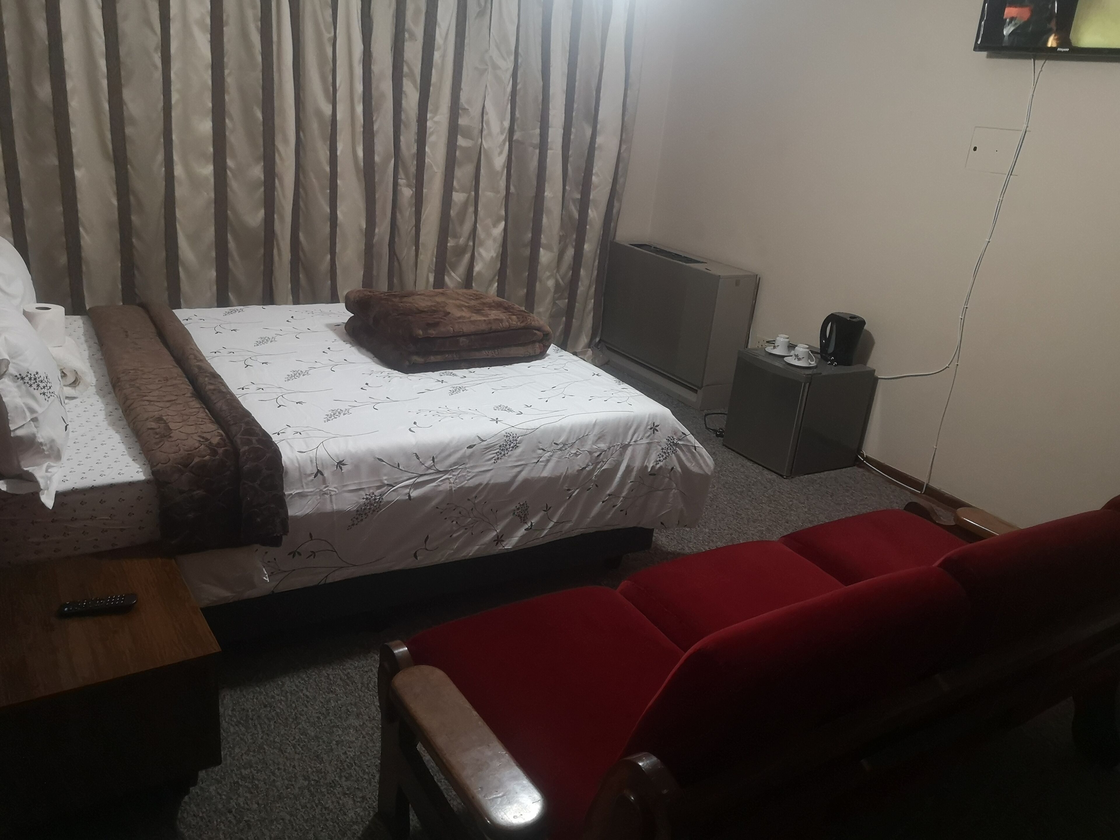 A hotel room with a bed, a TV, a small table, a red chair, and a curtain.