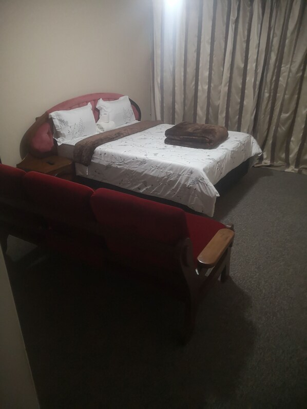 Un dormitorio con cama, sofá rojo, mesita de noche de madera y cortina.