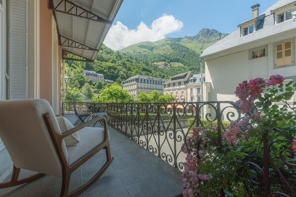 En balkon med en stol, gelænder i jern og udsigt over en by med bygninger og bjerge i baggrunden.
