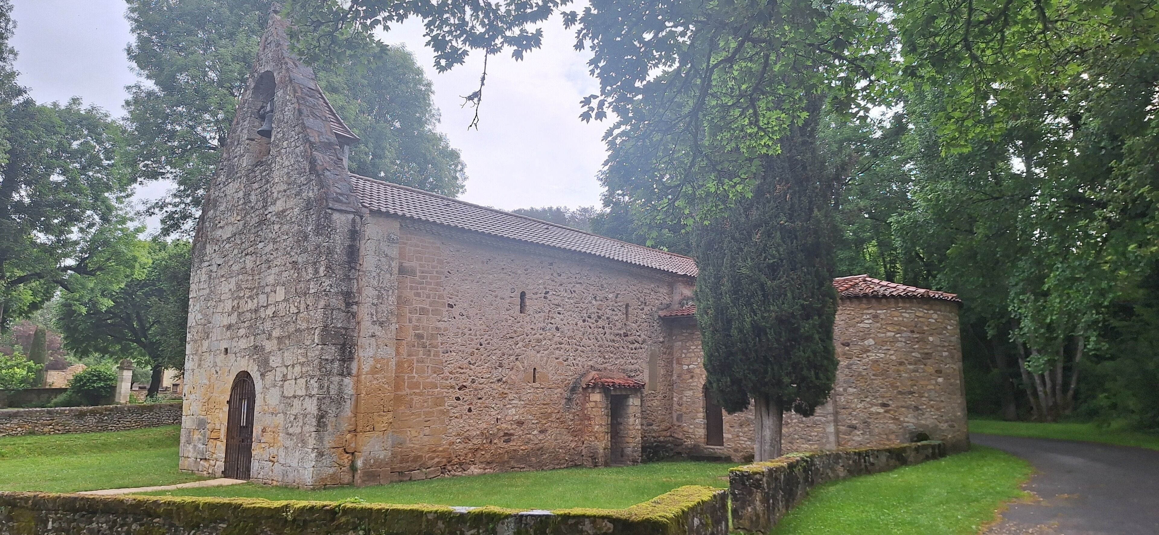 Un bâtiment en pierre, doté d’un toit en tuiles, d’une porte en arc de cercle et de fenêtres, le tout entouré de verdure.