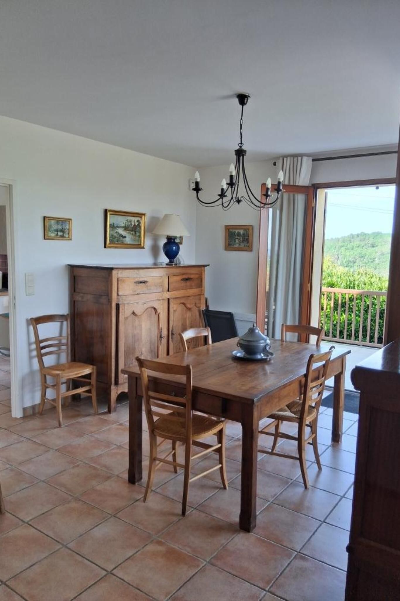 Un coin repas avec une table et des chaises en bois, un lustre et une vue sur la verdure par une fenêtre.
