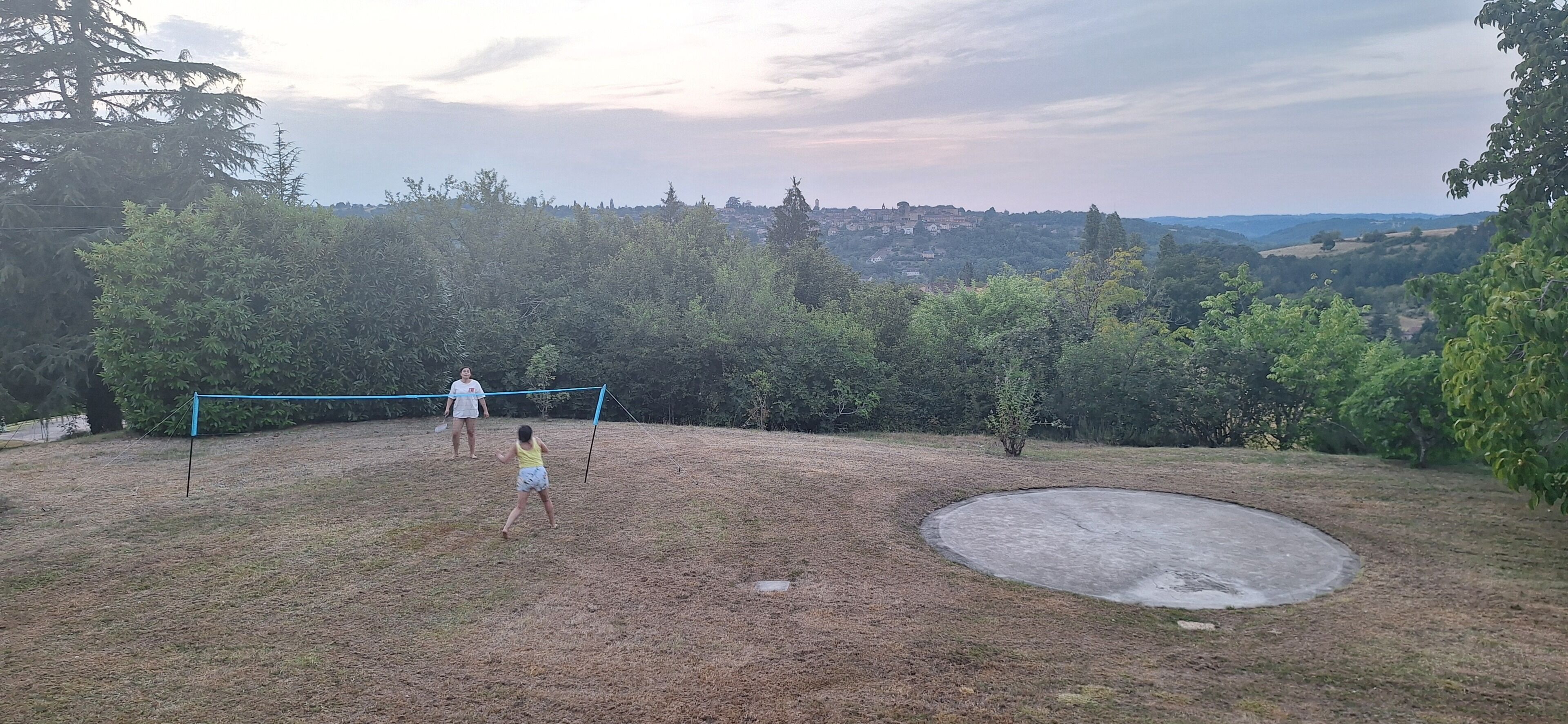An outdoor area with a volleyball net, a circular concrete feature, and a backdrop of trees and distant buildings.