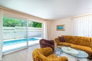 Ein Wohnzimmer mit Sofa, Sessel und Couchtisch, eine Glastür, die zu einem Pool führt, und einem Gemälde an der Wand.