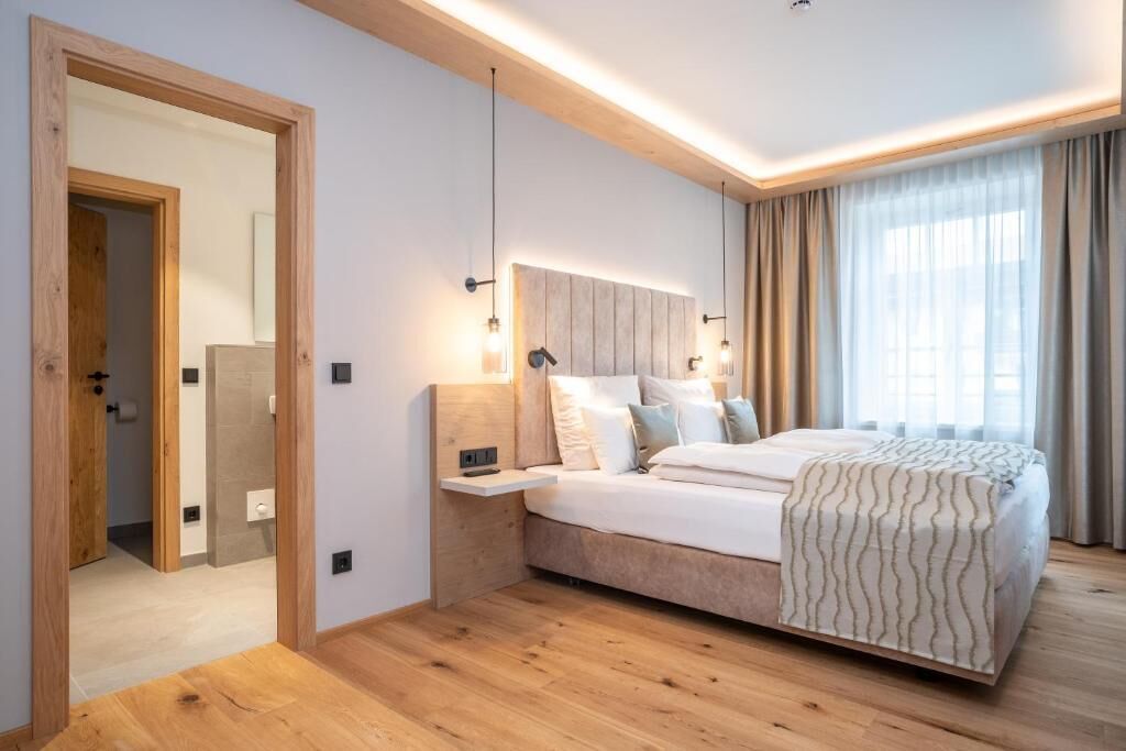 Un dormitori modern amb un terra de fusta, un llit amb llençols estampats, una nitzeta, una làmpada penjada a la paret i una finestra gran amb cortines.
