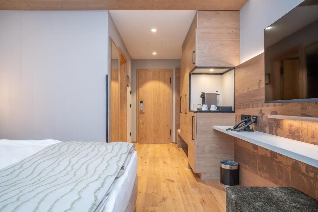 A modern hotel room with a bed, wooden flooring, a small seating area, a mirror, and a kitchenette.