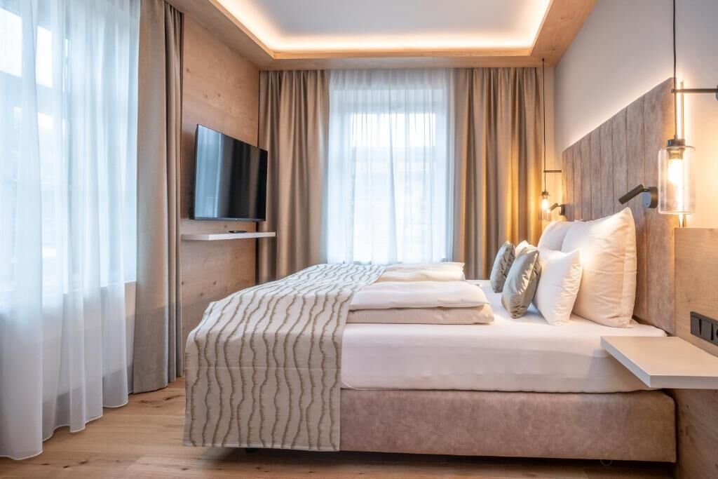 A modern hotel room with a bed, curtains, a mounted TV, and wall-mounted lighting.