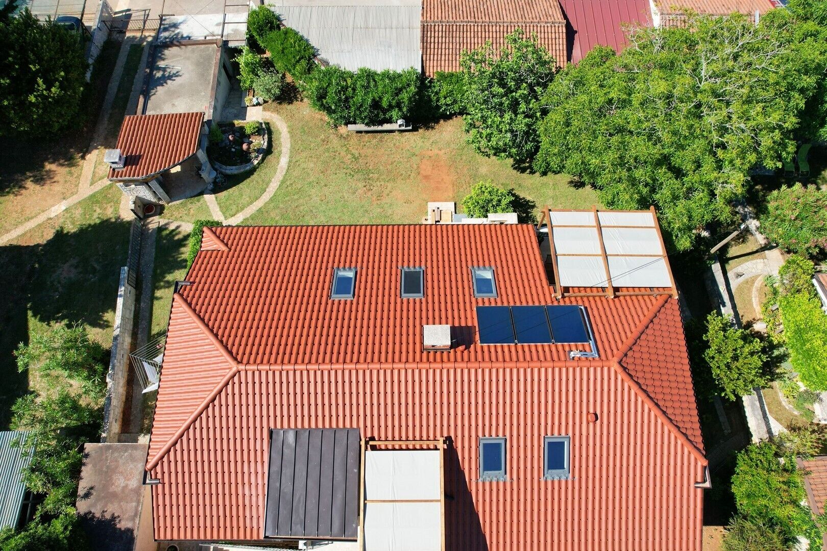 Ein mit roten Ziegeln gedecktes Dach mit Dachfenstern und Solaranlagen, umgeben von Grünflächen und angrenzenden Gebäuden.