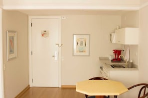 A room with a wooden floor, a white door, a small kitchen area with a red appliance, and a framed picture on the wall. - Apartment in Jardins with parking space and pool. (São Paulo)