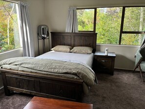 A bedroom with a wooden bed, two bedside tables, a nightstand, and a view of greenery through large windows. - A Piece of Paradise With Your own Entrance! (Auckland)