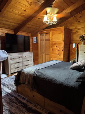 A wooden cabin bedroom with a bed, dresser, TV, and a fan.