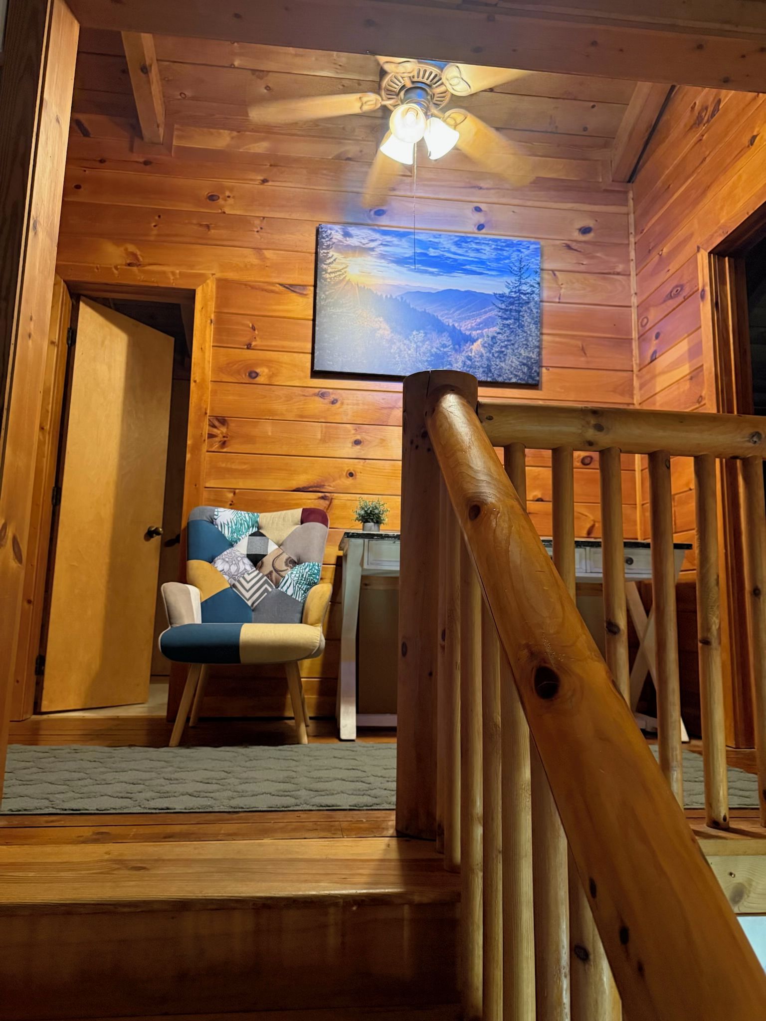 Una escalera de madera con barandilla que conduce a una habitación con ventilador de techo y un cuadro en la pared.