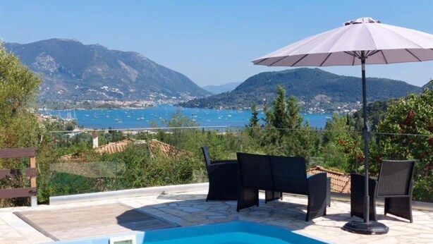 Piscina con mobiliario exterior y una sombrilla, con vistas a un paisaje costero con montañas y una masa de agua.
