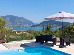 A swimming pool with outdoor furniture and an umbrella, overlooking a coastal landscape with mountains and a body of water. - Villa Avlaki (Vlikhon)