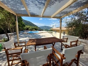 A covered outdoor seating area with a wooden table and chairs, overlooking a pool and a mountainous landscape. - Villa Avlaki (Vlikhon)