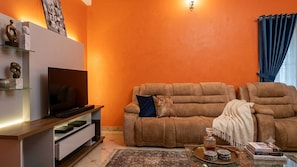 Una sala de estar con un sofá, un televisor de pantalla plana, una mesa de centro con libros y una bandeja, y una pared decorada con una obra de arte con textura.