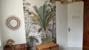 A bedroom with a bed, a chair, a small table, a wicker chest, a wall mural, and a door. - Large, single-story, air-conditioned house with 5 bedrooms near the Marais Poitevin (Saint-Pierre-d'Amilly)