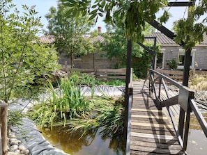 Une terrasse en bois surplombe un étang, entourée de verdure, et un mur de pierres se dresse en arrière-plan.