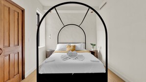 Quarto com cama sob um arco preto, roupa de cama branca, uma mesa de cabeceira de madeira, uma planta em vaso e uma porta de madeira.