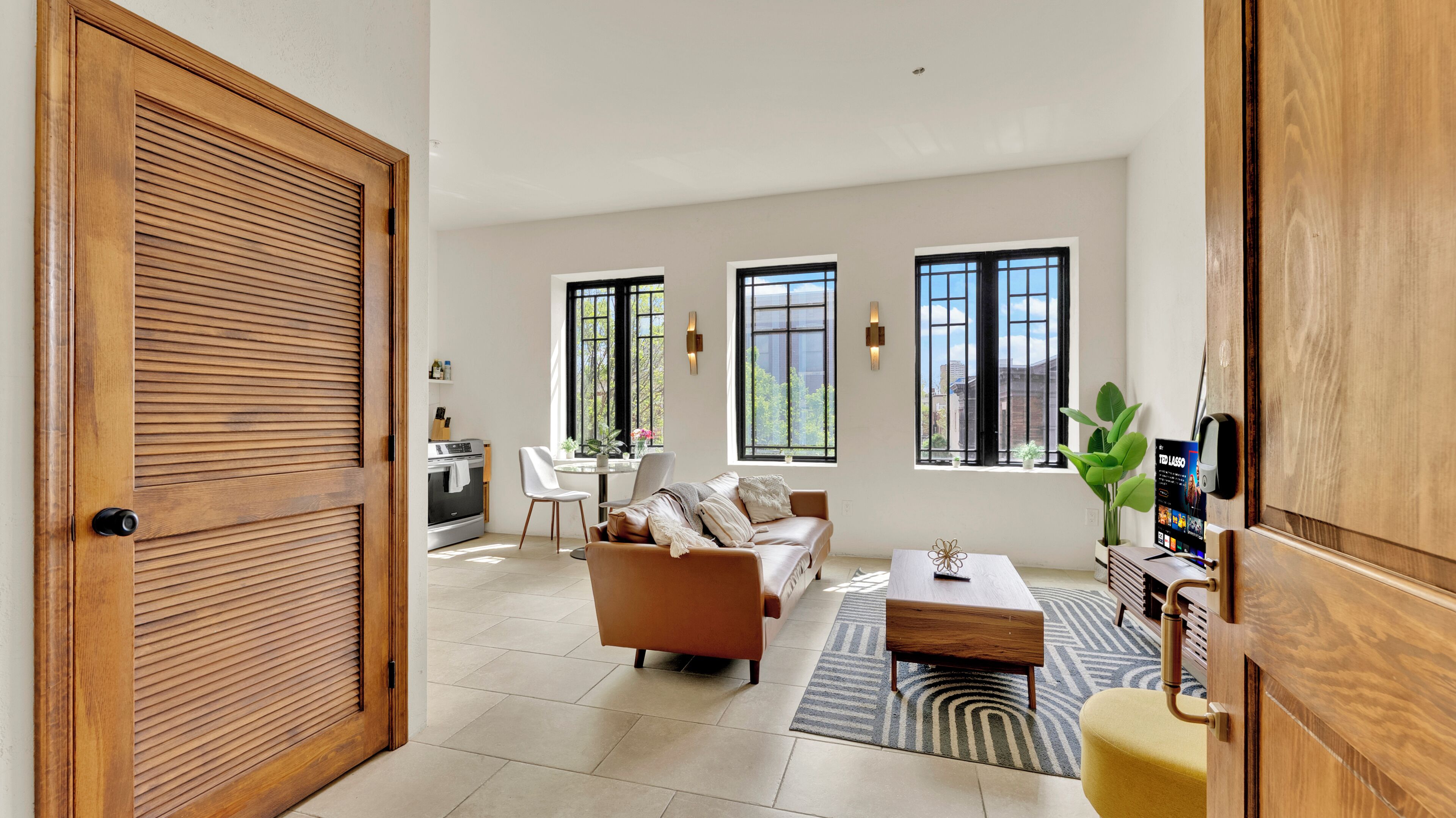 A spacious living area with a sofa, coffee table, and a dining table with chairs. There are large windows providing natural light and views of the outside.