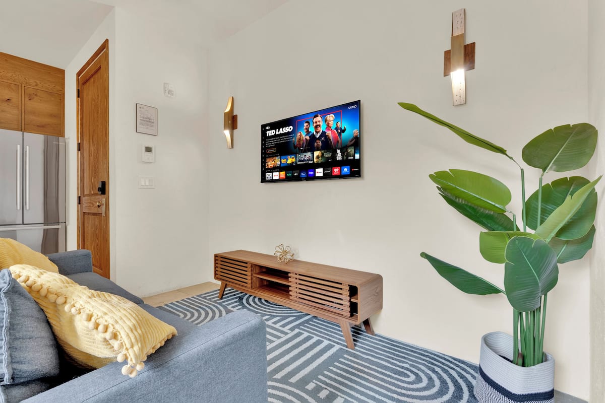 A living room with a sofa, a wooden TV stand, a potted plant, and a kitchen area with a refrigerator and cabinets.