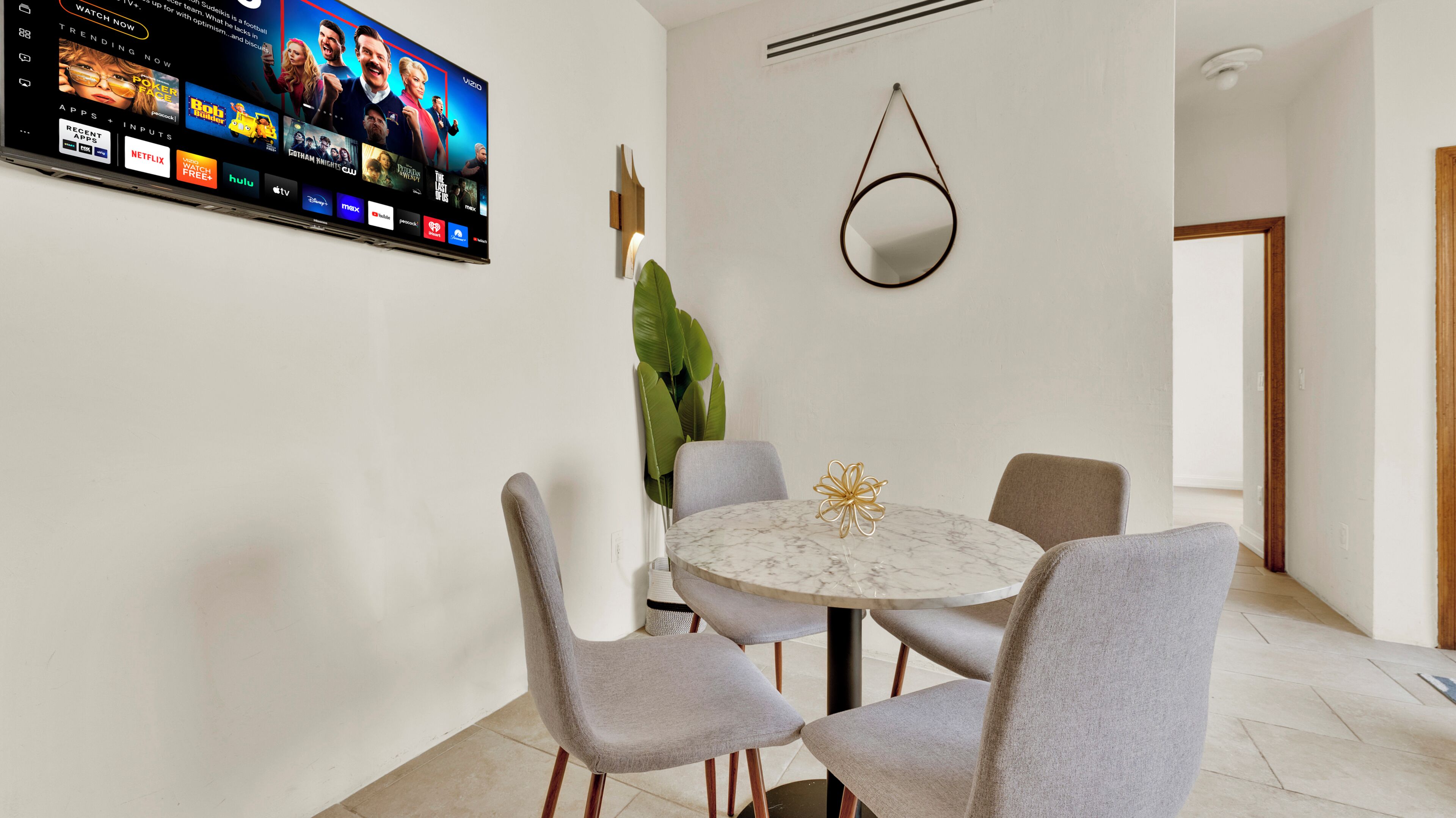 Ein moderner Essbereich mit Fernseher, der Streaming-Dienste anzeigt, einem runden Marmortisch und grau gepolsterten Stühlen.