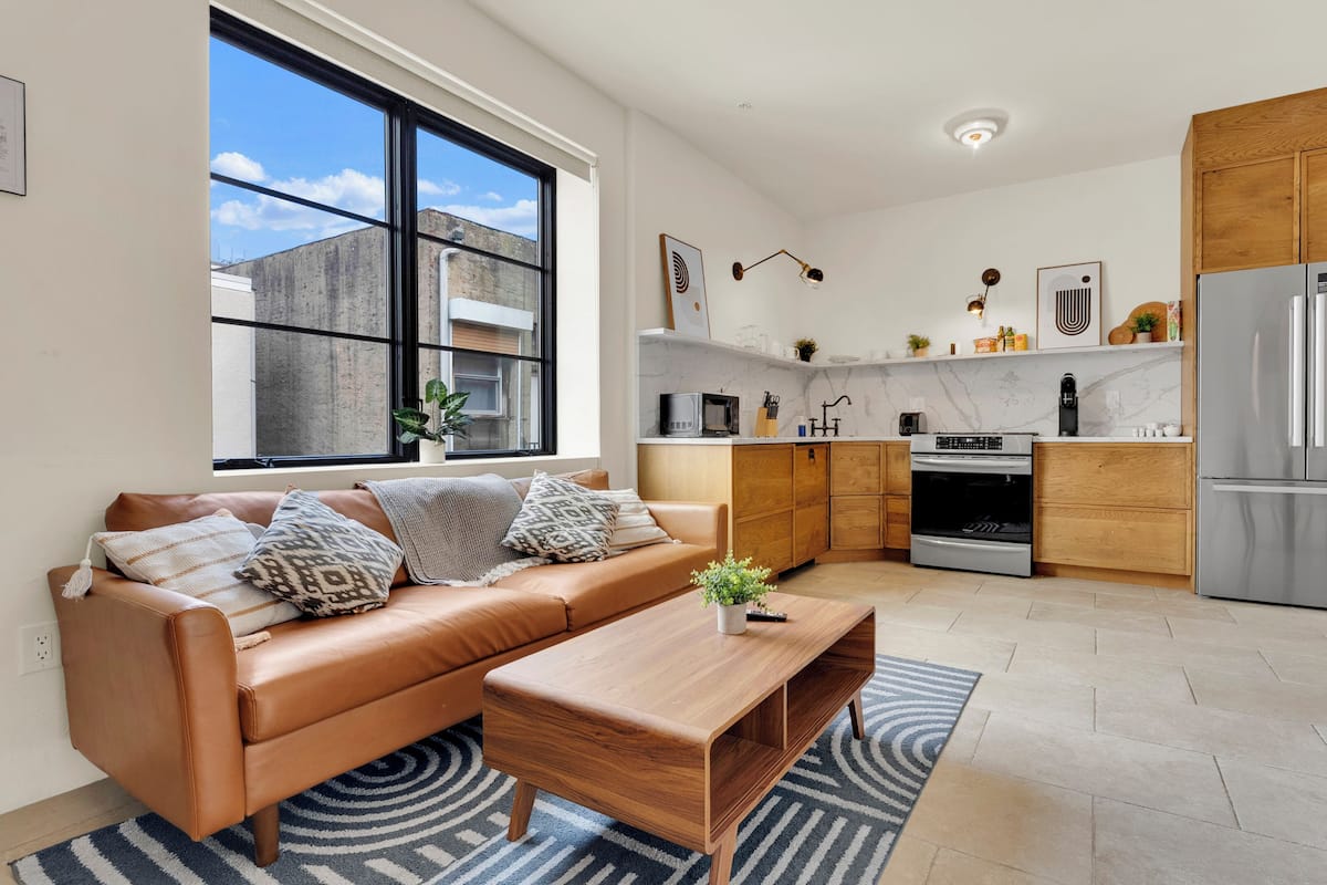 A modern apartment with a leather sofa, wooden coffee table, kitchen with stainless steel appliances, and a large window.
